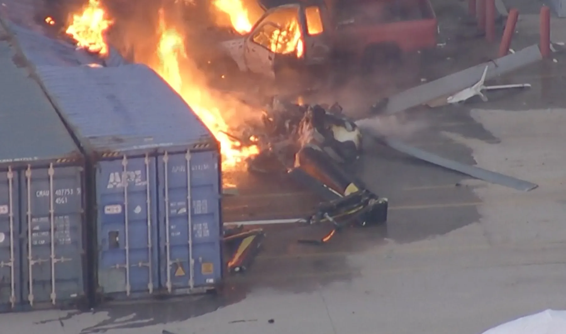 Burning helicopter wreckage lies beside shipping containers and vehicles on a paved surface.