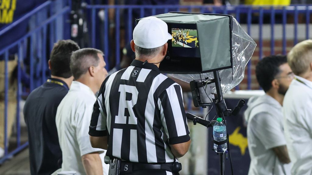 Football referee reviews a replay monitor beside the sideline during a game.