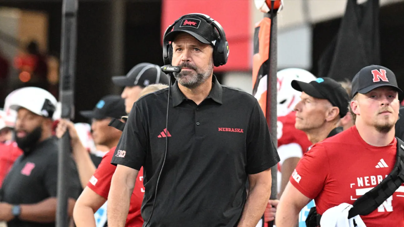 Nebraska football coach wearing a headset stands on the sideline among players and staff