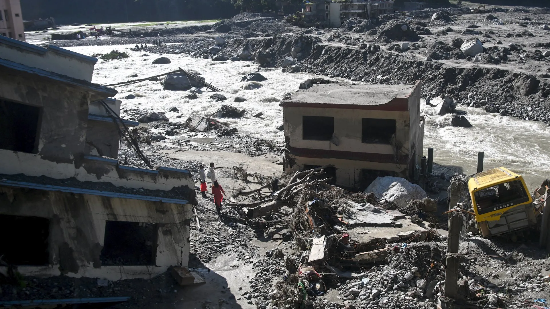 Floodwater rushes past damaged buildings, rubble, and a partly buried yellow truck as two people walk through debris.