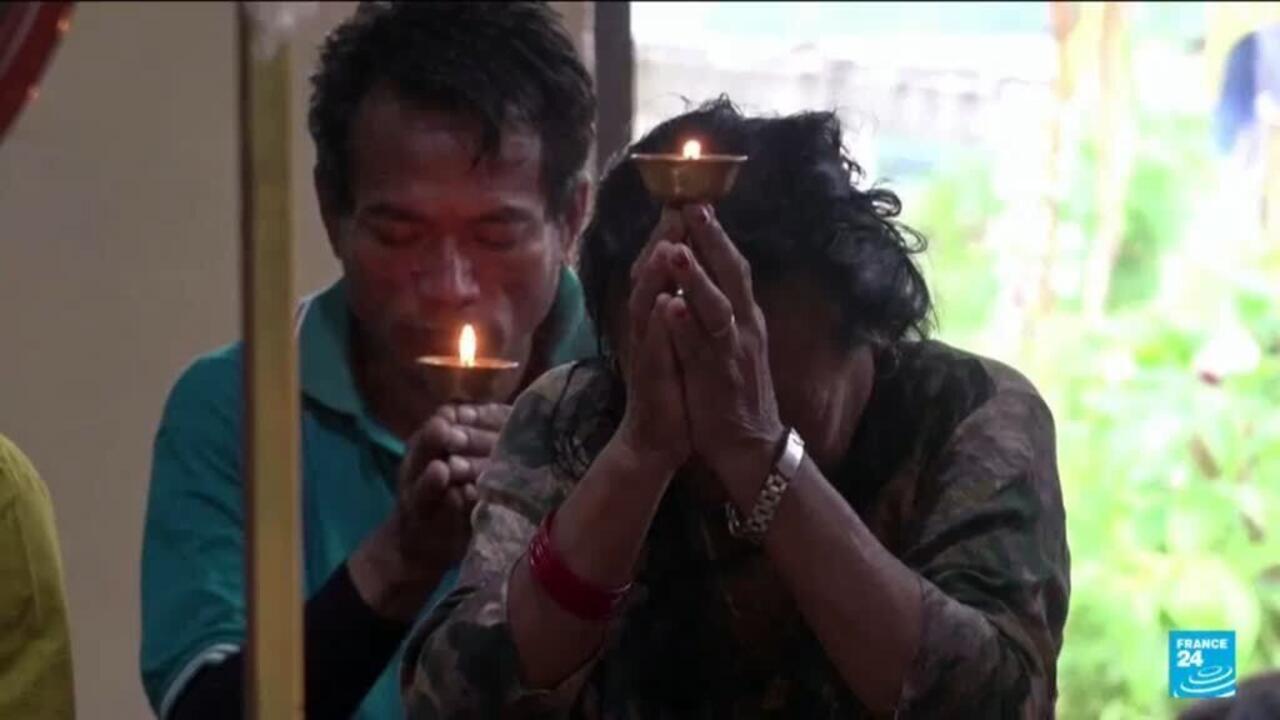 Two people hold lit oil lamps with their hands clasped in prayer indoors.