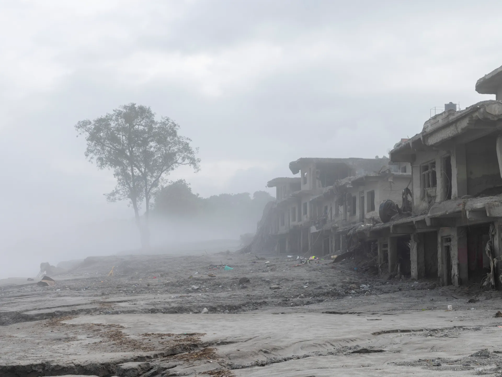 Fog shrouds a muddy, debris-strewn settlement beside severely damaged buildings and a lone tree.