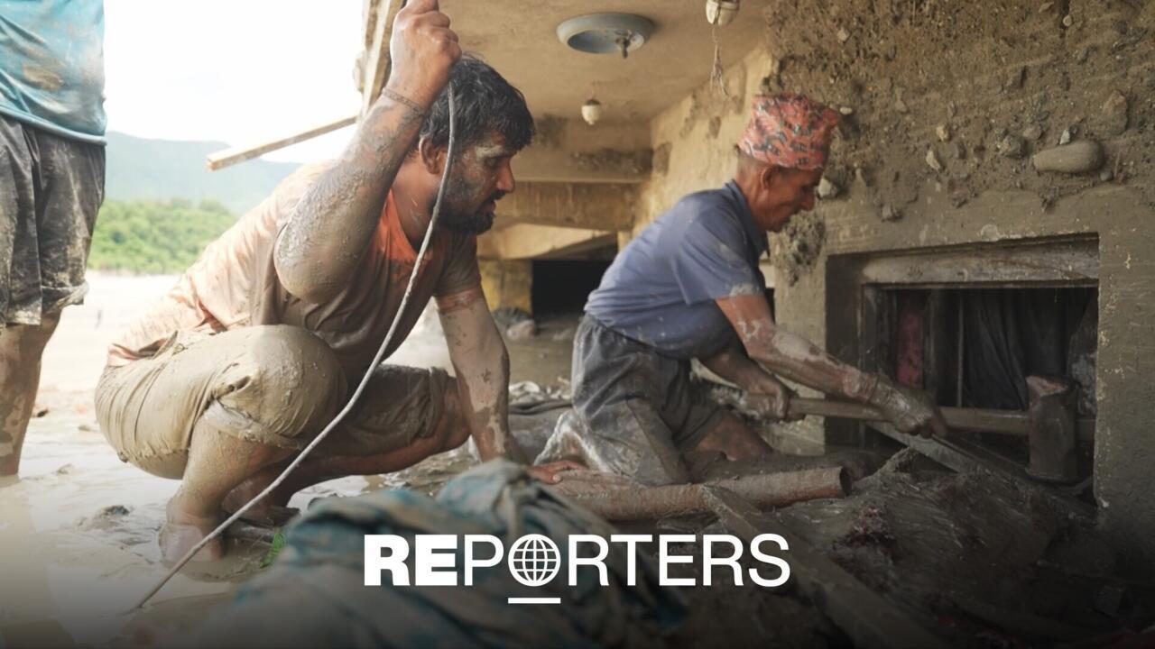 Mud-covered workers repair a damaged building beside floodwater, with tools and debris visible.