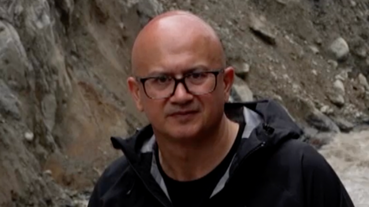 Bald man wearing glasses and a dark jacket stands beside a rocky excavation