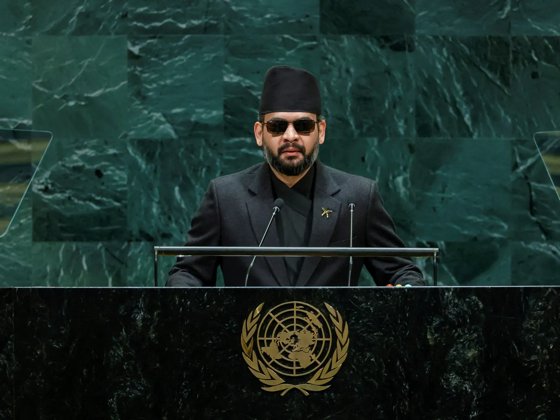 A bearded man in a black suit and cap speaks at a lectern before a dark green backdrop.