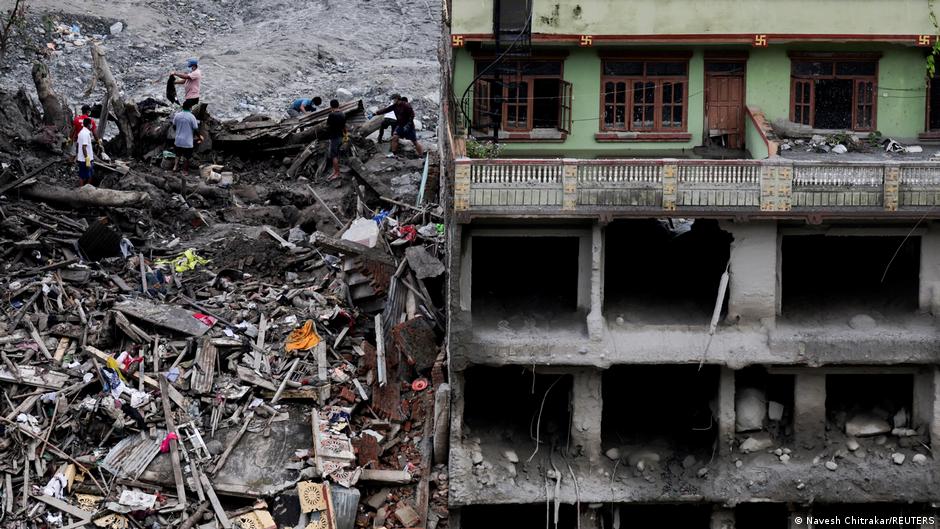 People search rubble beside a severely damaged multi-story building with exposed interiors and debris.