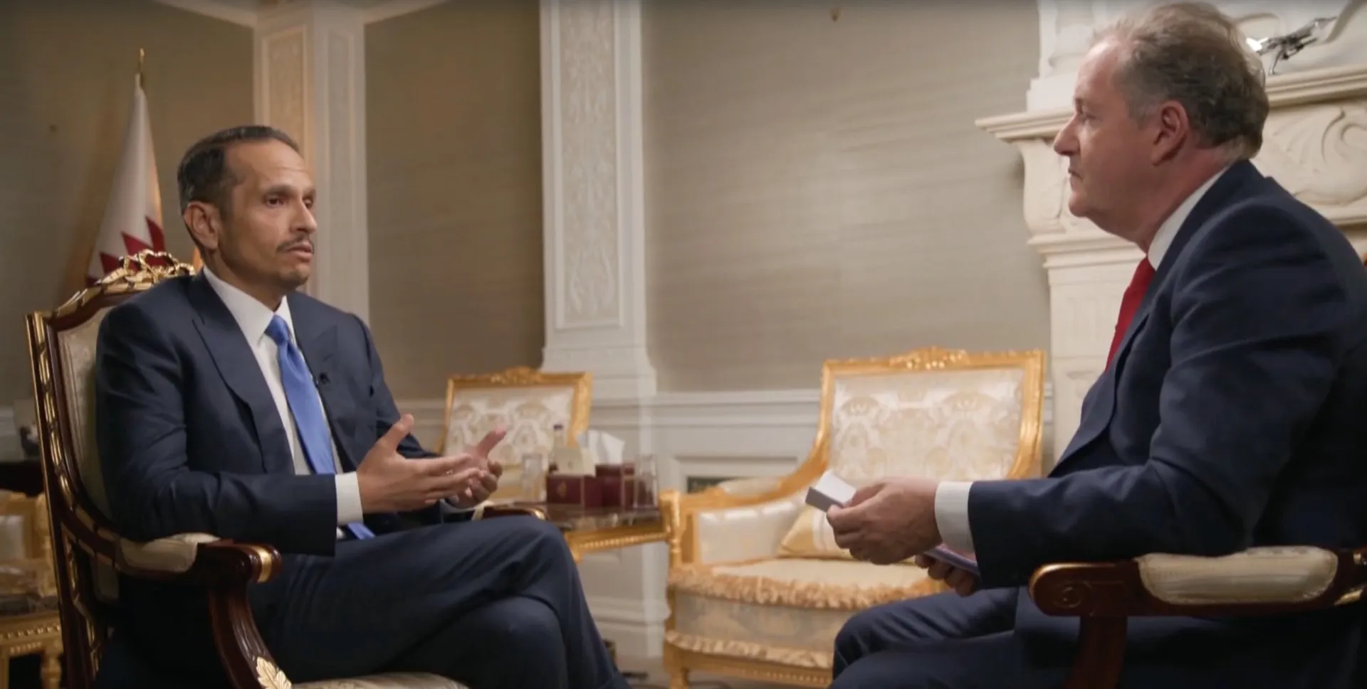Two suited men sit facing each other in an ornate room during an interview.