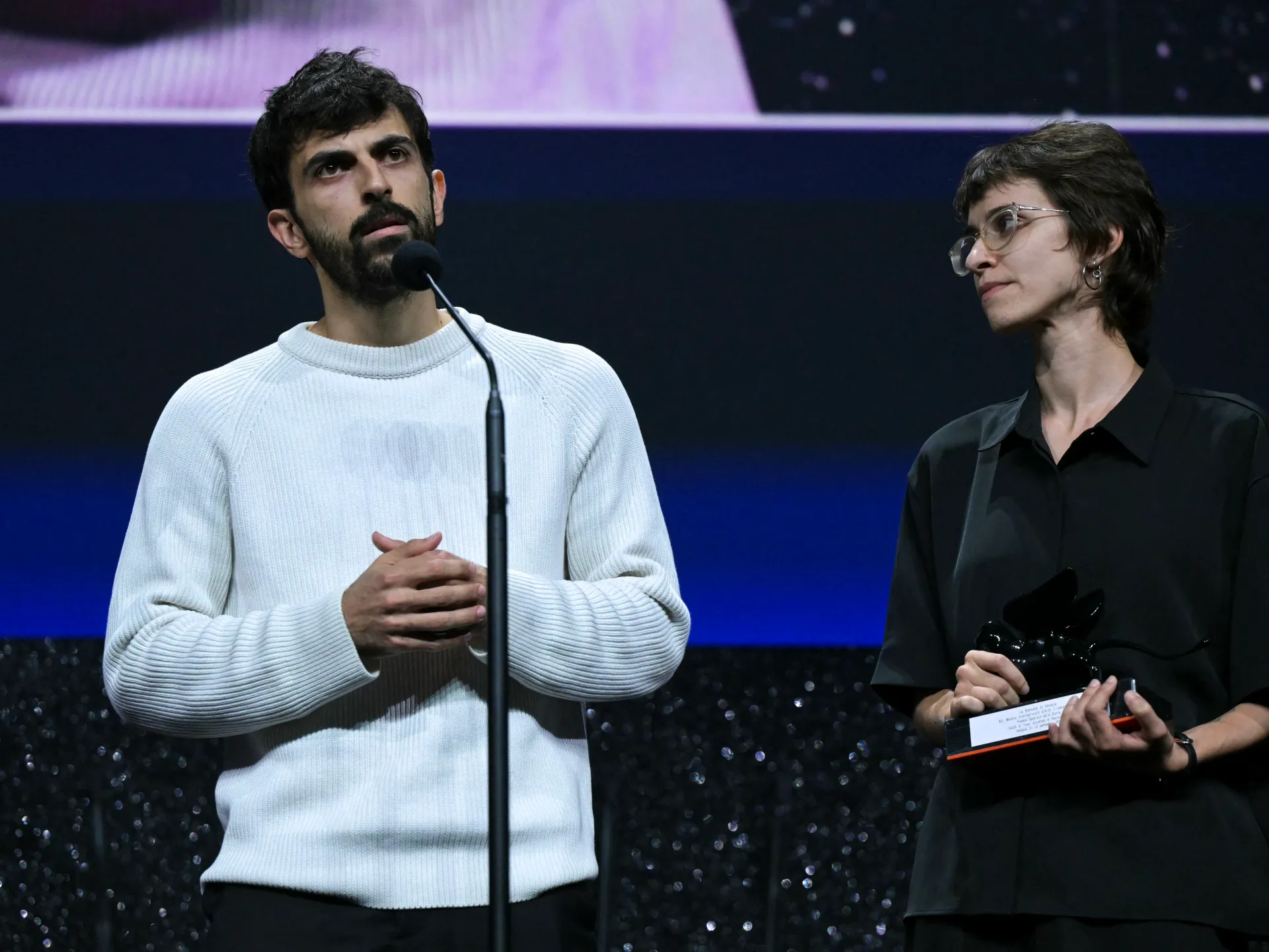 A man speaks into a microphone beside a woman holding a camera on a dark stage.