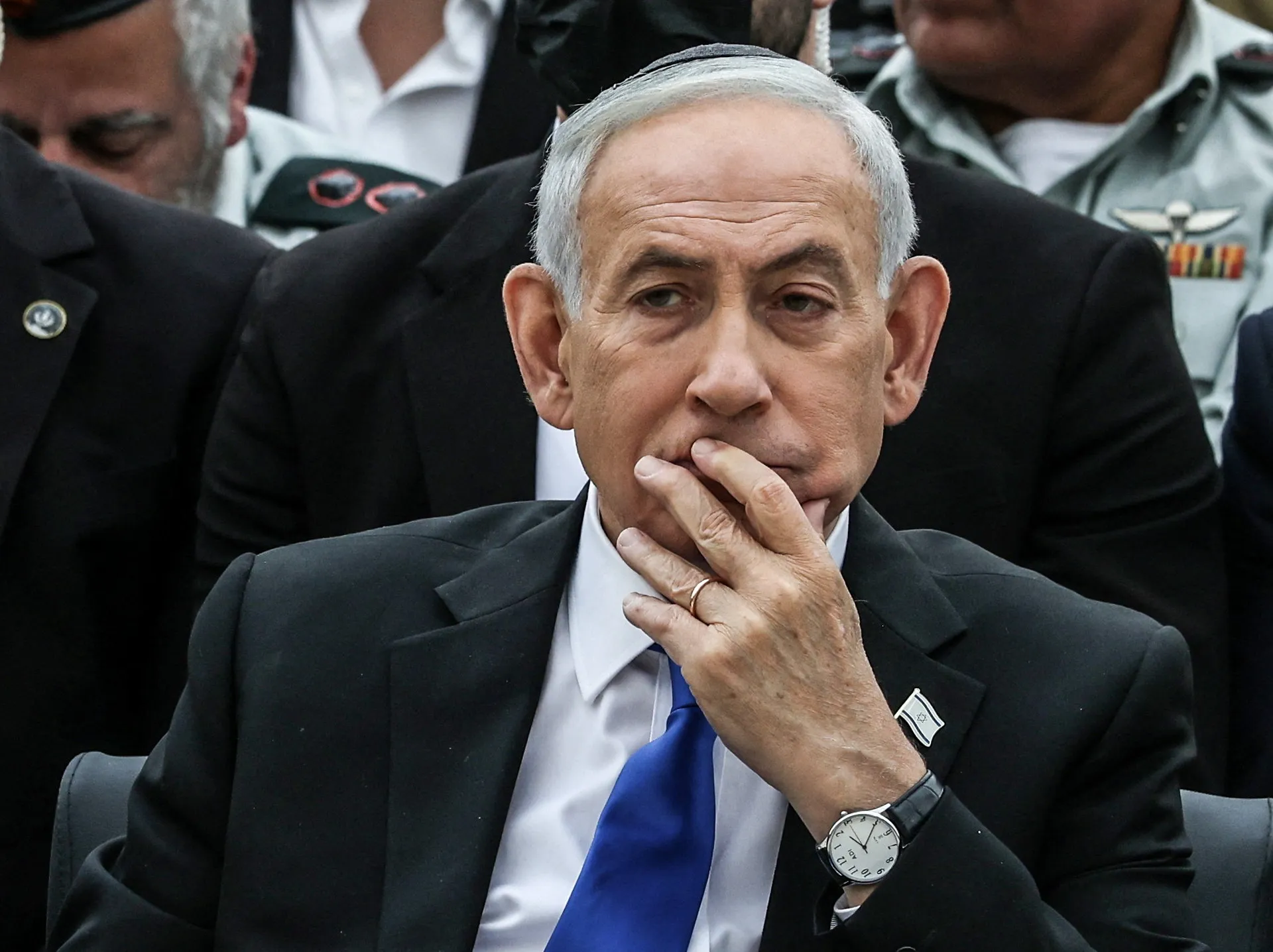 Gray-haired man in suit rests his hand on his chin while seated among uniformed officials.