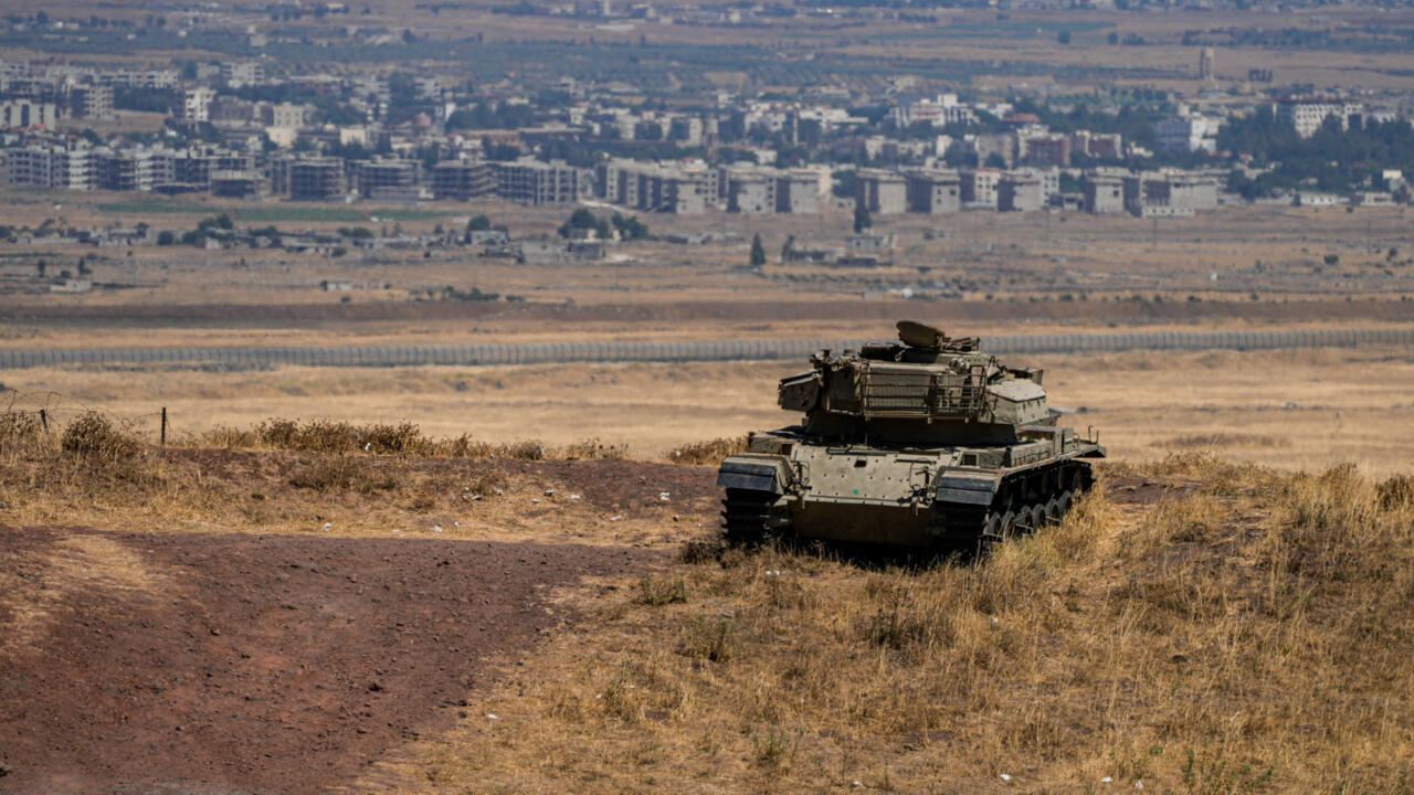Armored tank positioned on a dry hillside overlooking fields and a densely built town