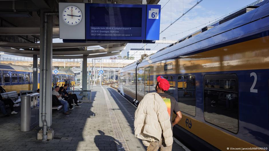 Passengers wait beside a blue and yellow train at a station platform under electronic departure boards.
