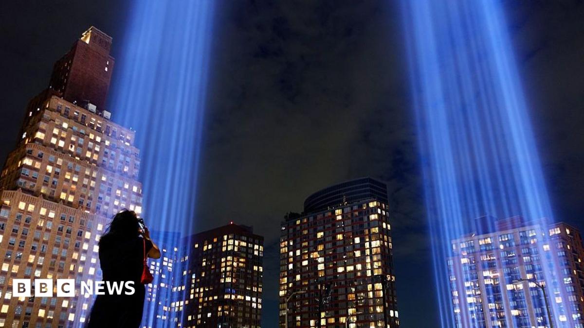 Blue memorial light beams rise between illuminated skyscrapers at night as a person photographs them.