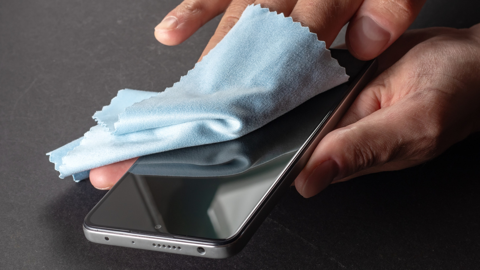 Person uses a soft blue cloth to clean a smartphone screen on a dark surface