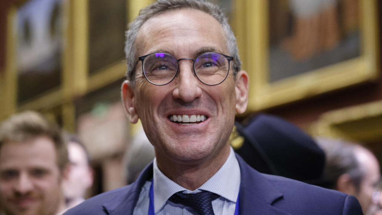 Smiling gray-haired man wearing round glasses and a suit at an indoor gathering