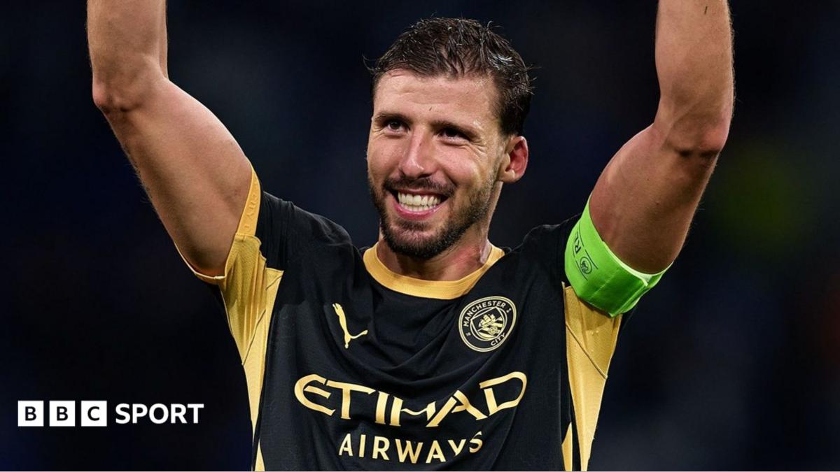 Manchester City footballer in black and gold kit raises both arms, wearing a green captain’s armband
