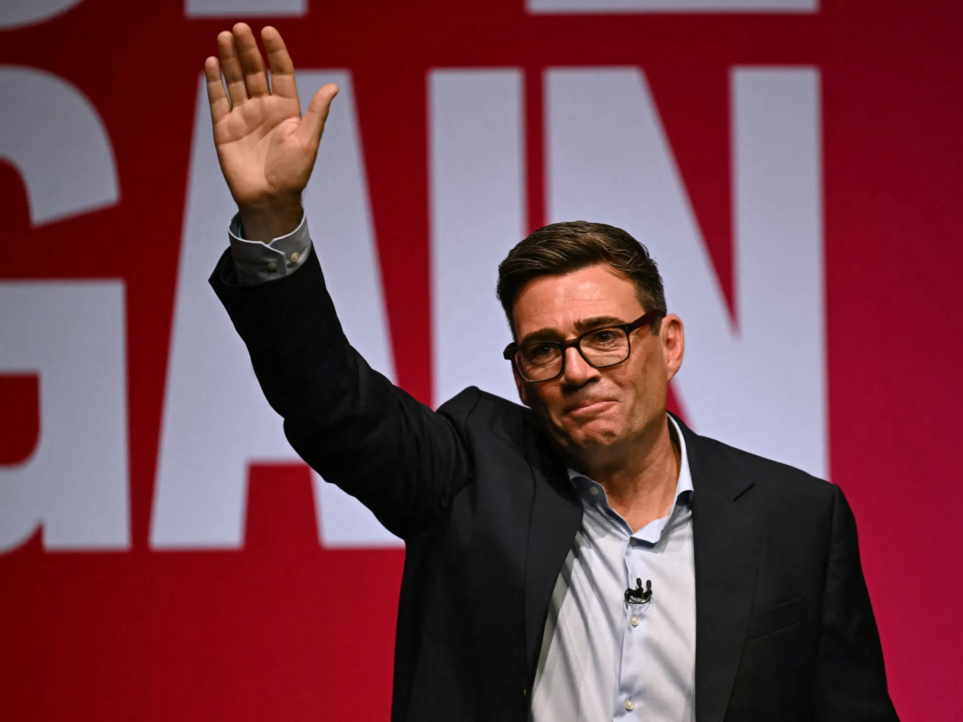 A man in glasses and a dark suit raises one hand before a red backdrop with large white lettering.