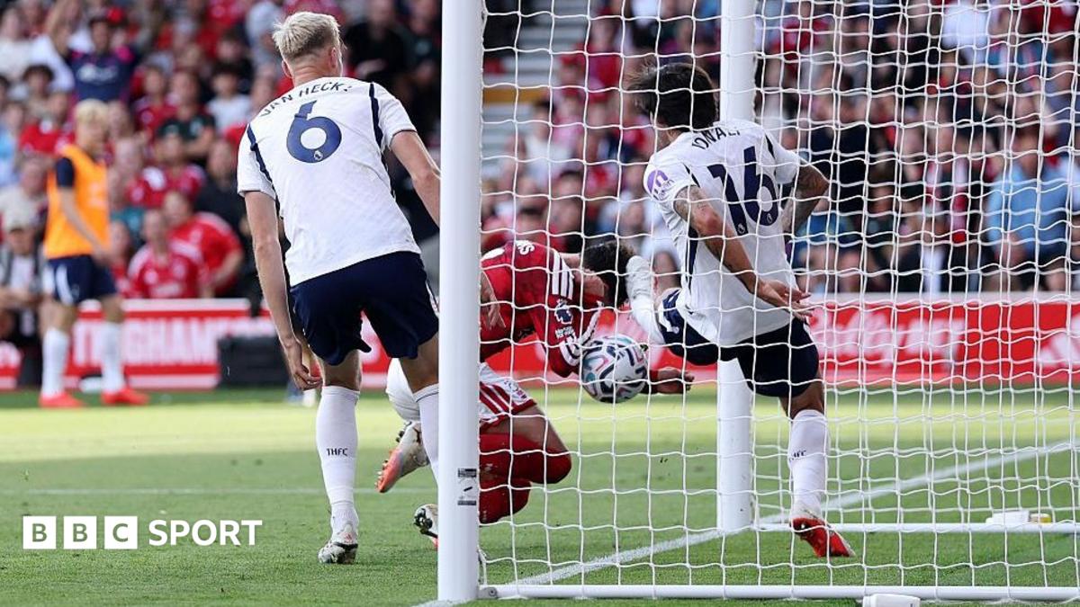 Footballers in white and red contest the ball near the goal, viewed through the net.