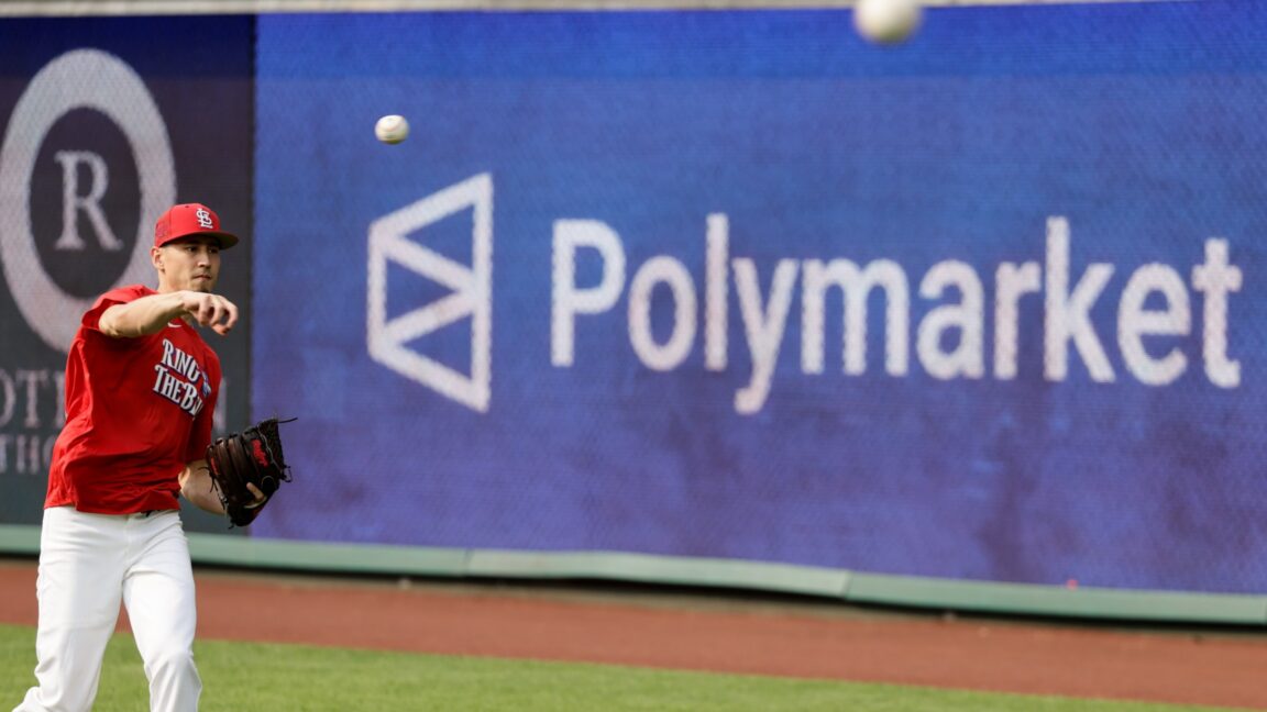 Baseball player in a red uniform swings as a screen behind him displays the word Polymarket.