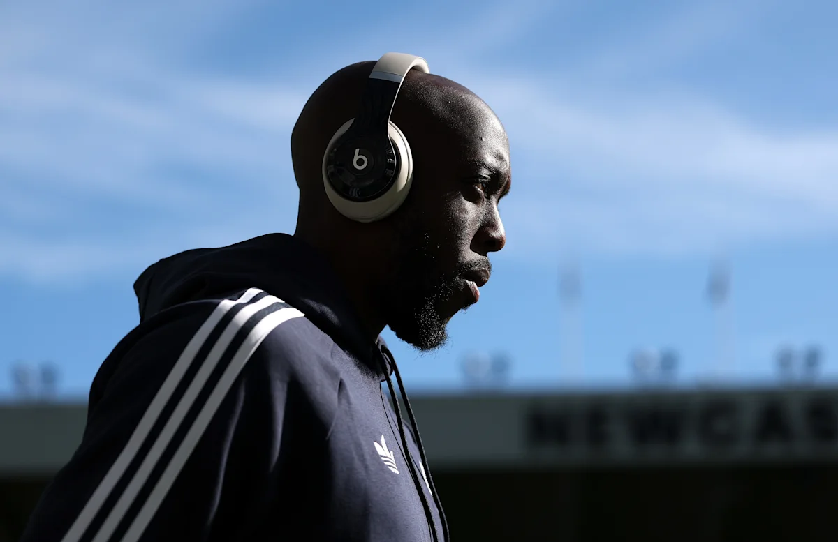 Bald man wearing headphones and a dark Adidas jacket walks past a football stadium