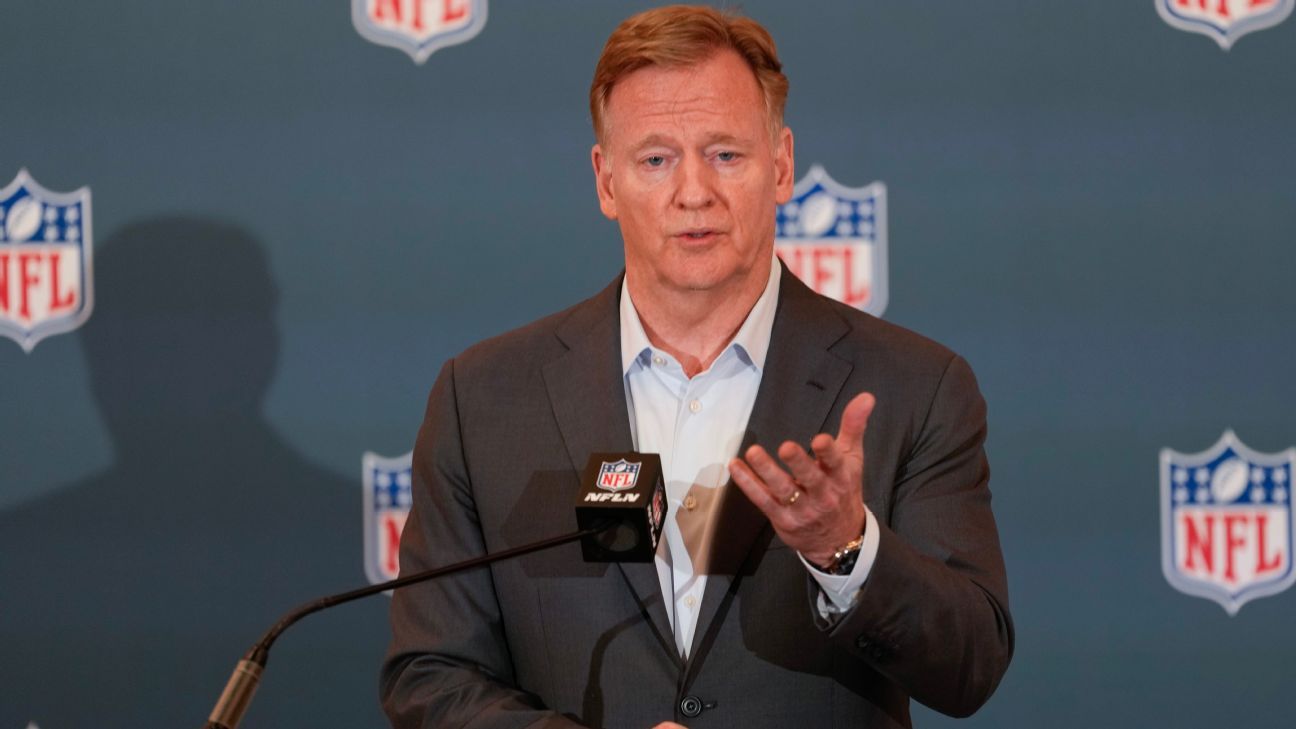 Man in suit gestures while speaking at a podium before an NFL-branded backdrop