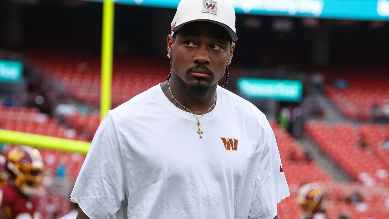 NFL player wearing a white Washington shirt and cap stands on a football field.