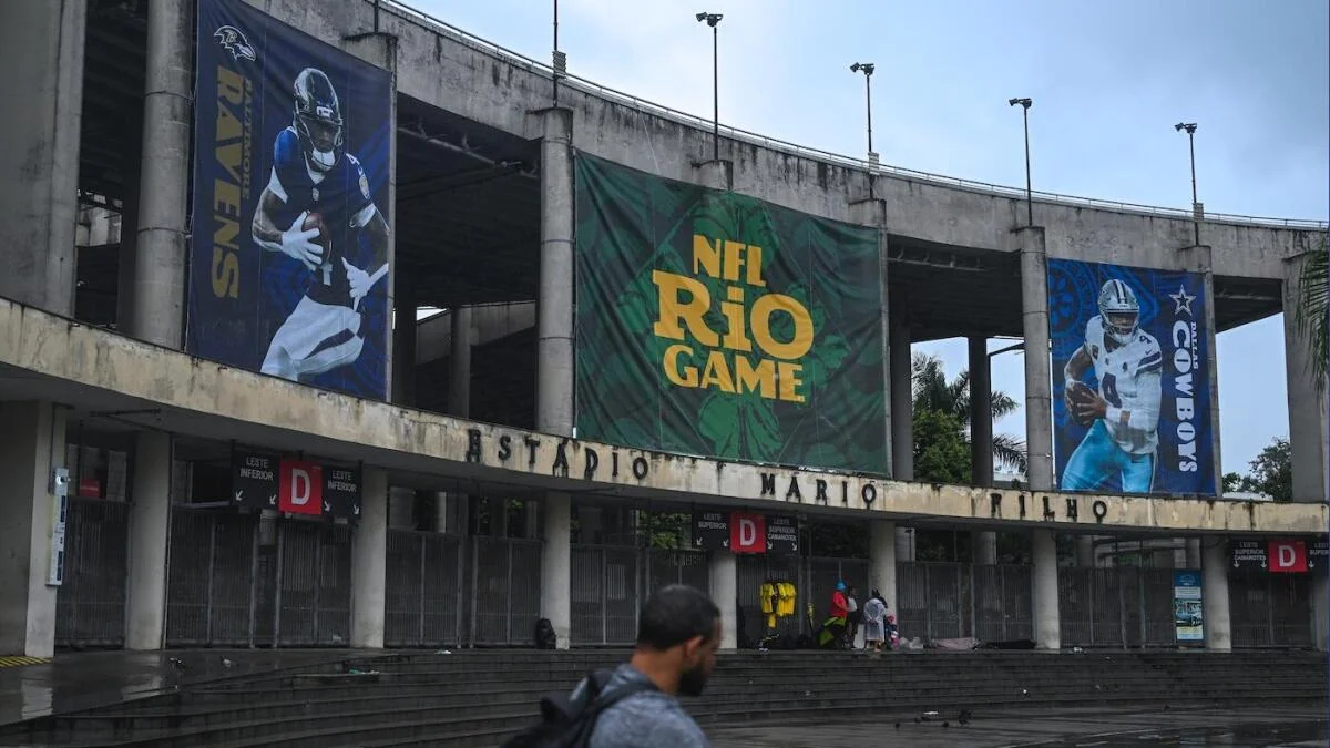 Concrete stadium facade displays NFL Rio Game and Ravens and Cowboys banners above wet entrance steps.