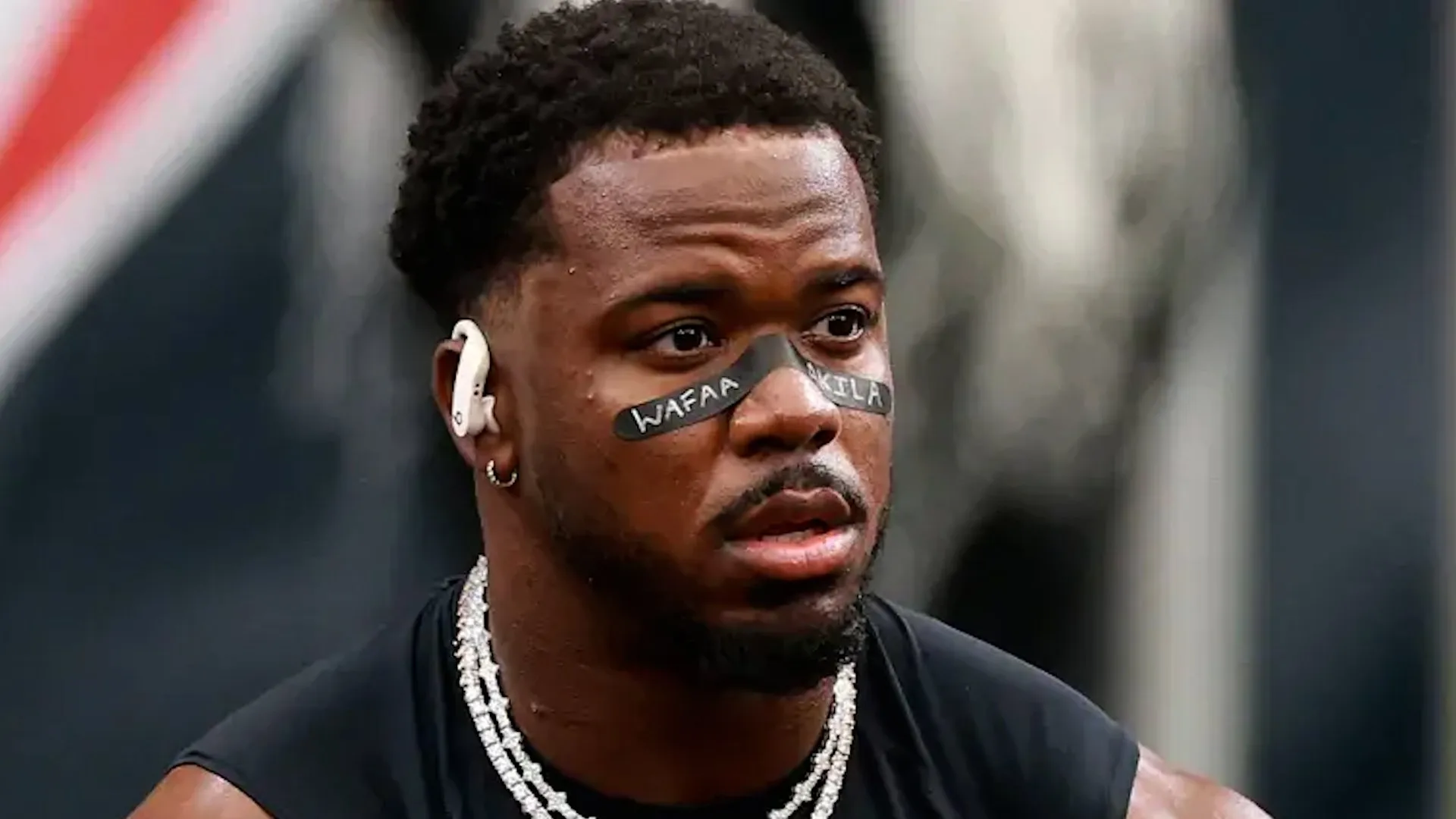 Football player wearing black eye paint, white earbuds, layered chains, and a sleeveless black shirt