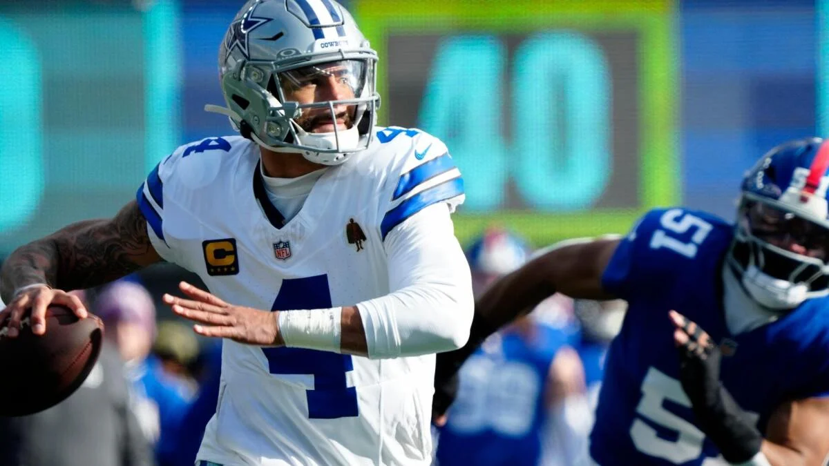 Dallas Cowboys quarterback in white uniform prepares to throw as a New York Giants defender pursues.