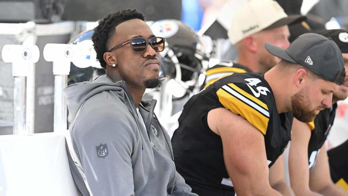 A man in a gray hoodie and sunglasses sits beside Steelers players on the sideline.