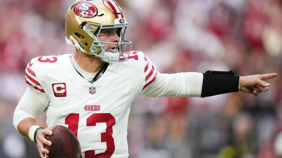 San Francisco 49ers quarterback in helmet points downfield while holding a football.