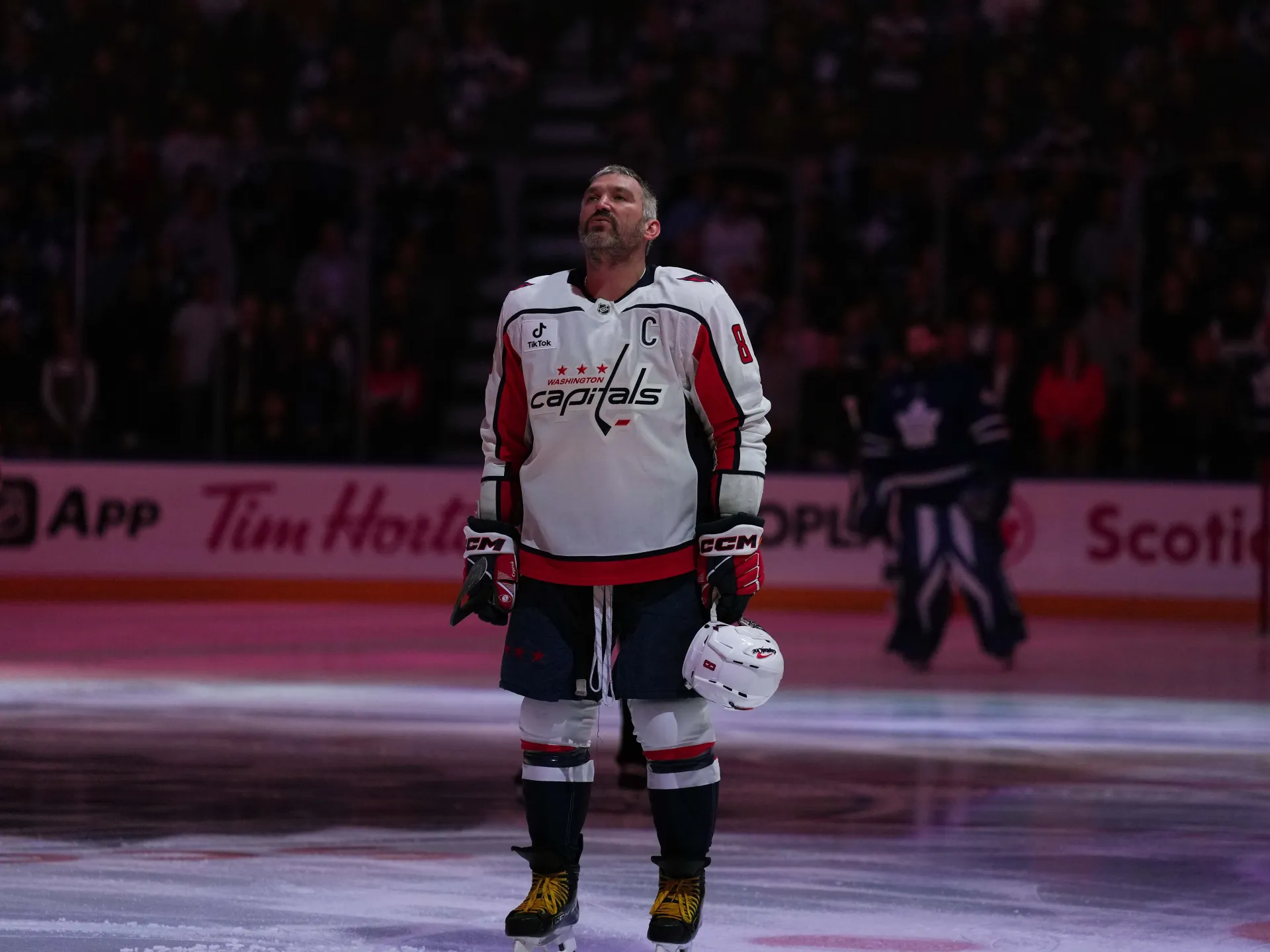 Hockey player in Washington Capitals uniform stands on ice holding his helmet during a game.