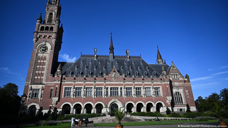 Historic red-brick Gothic building with clock tower, arches, spires, and landscaped grounds under blue sky