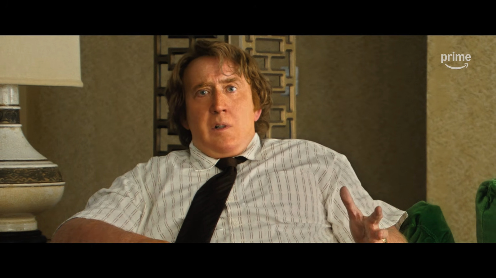 A man in a patterned white shirt and black tie gestures while seated indoors.