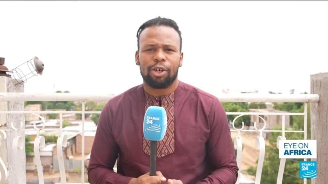 Man in maroon traditional clothing holds a France 24 microphone on a rooftop terrace.