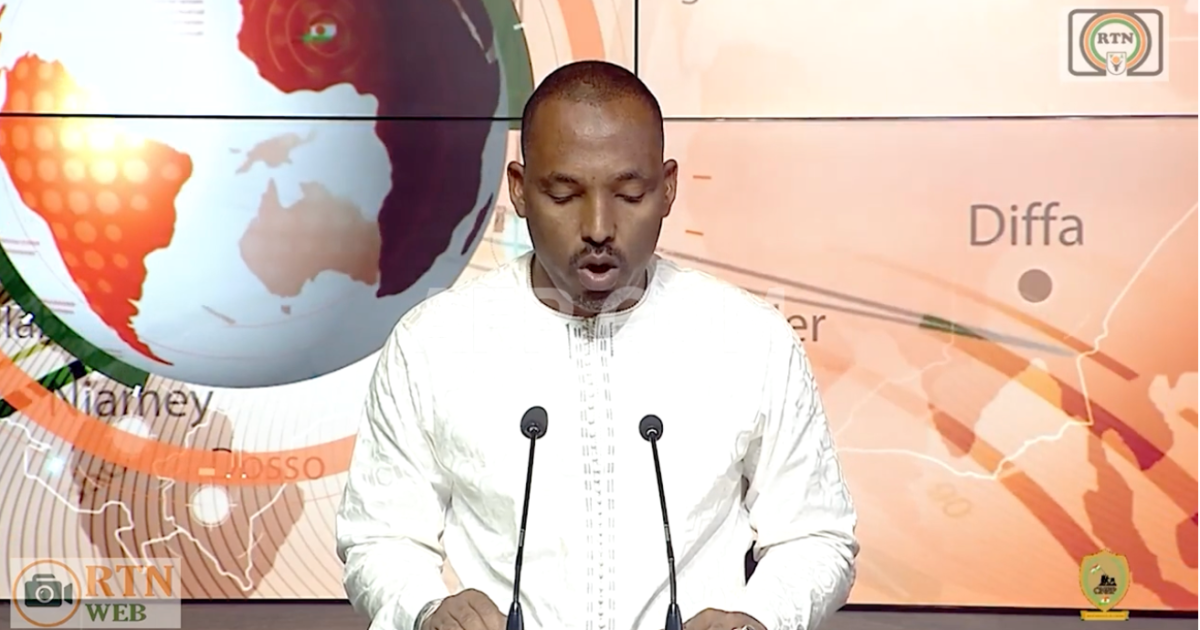 Man in white traditional attire speaks at a podium with microphones before a televised news backdrop.