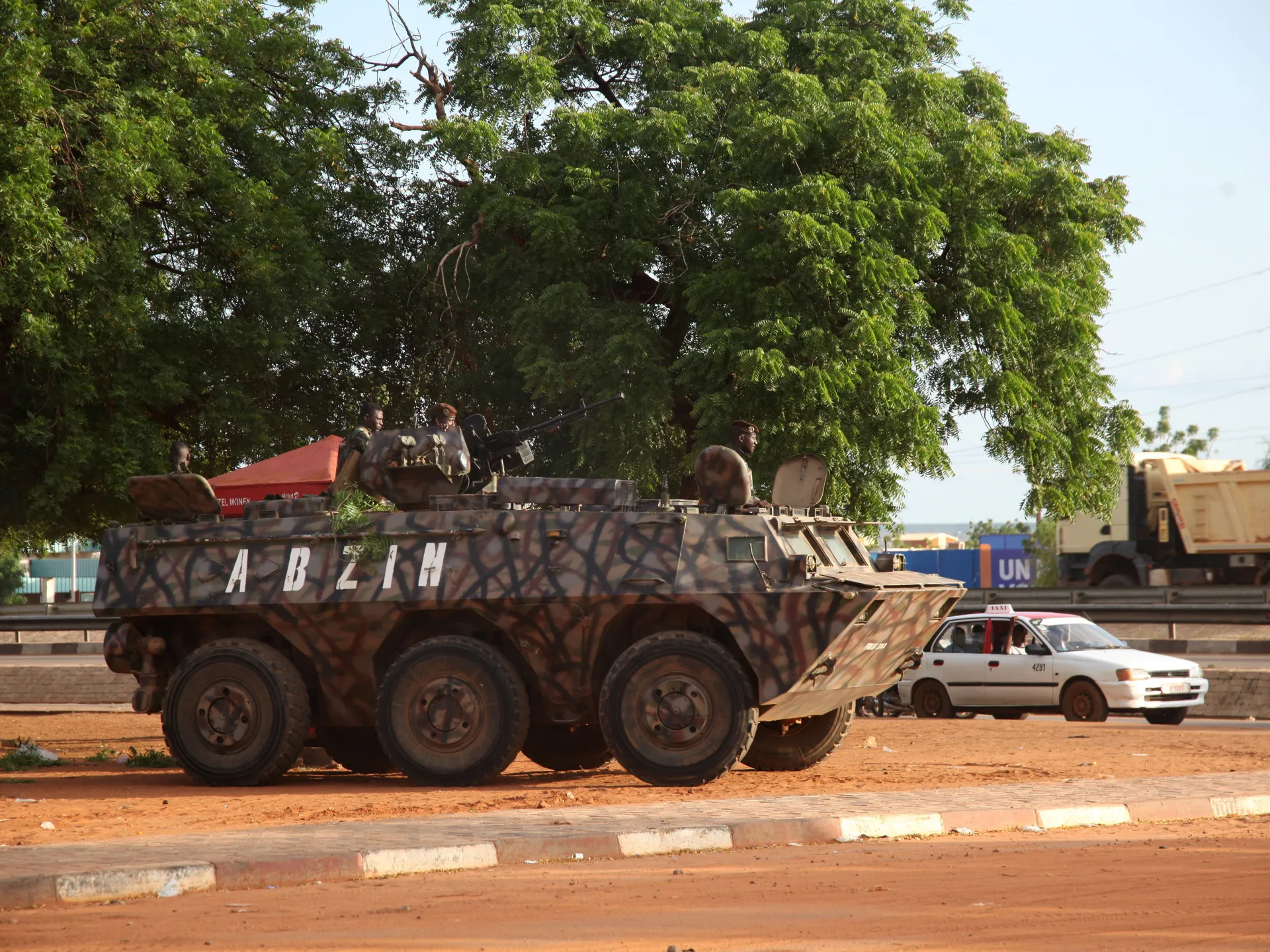 Camouflaged armored personnel carrier with soldiers parked beside a roadway under large trees
