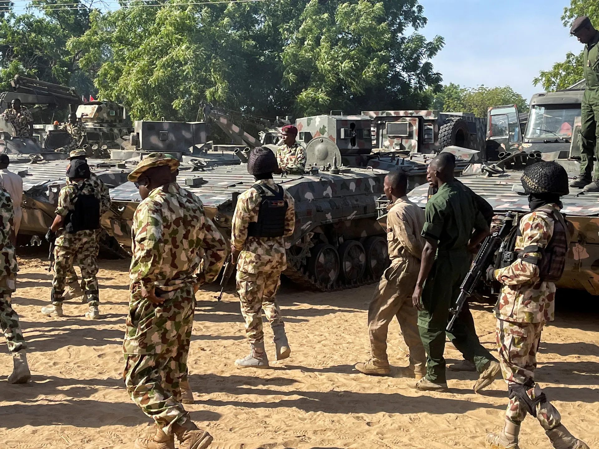 Soldiers in camouflage uniforms stand beside armored vehicles in a sunlit sandy area.