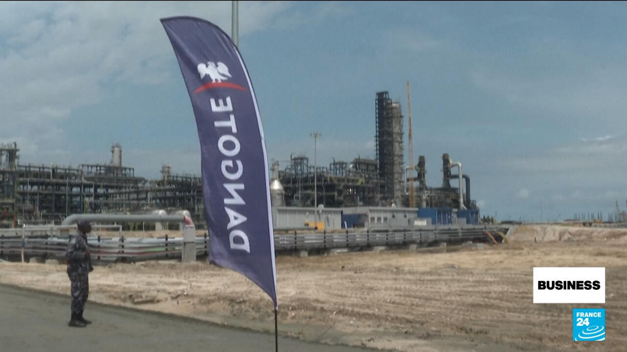 Dangote flag stands before an industrial refinery, with a worker beside roadside piping.