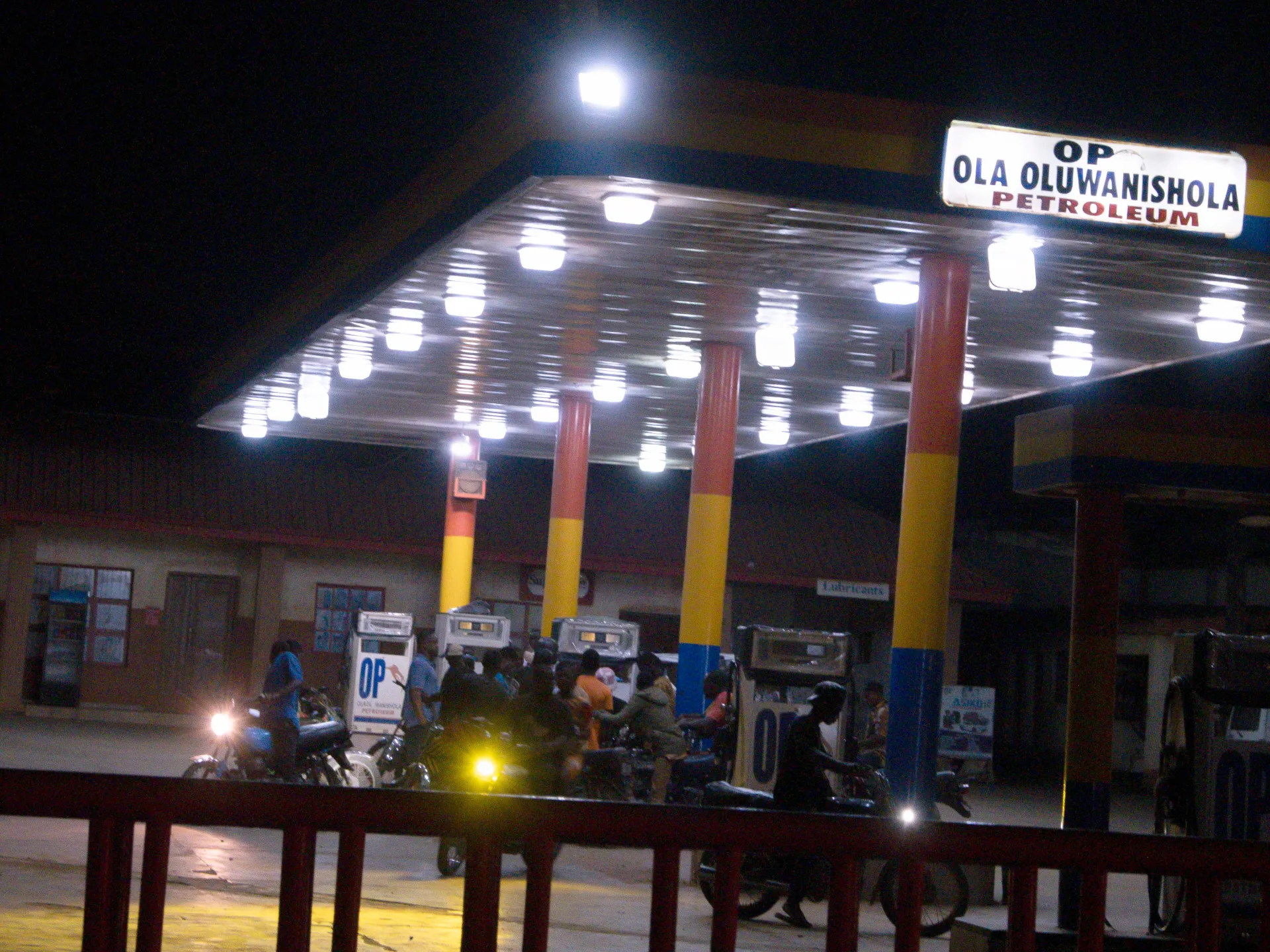 A crowded petrol station at night, with motorcycles beneath a brightly lit canopy.