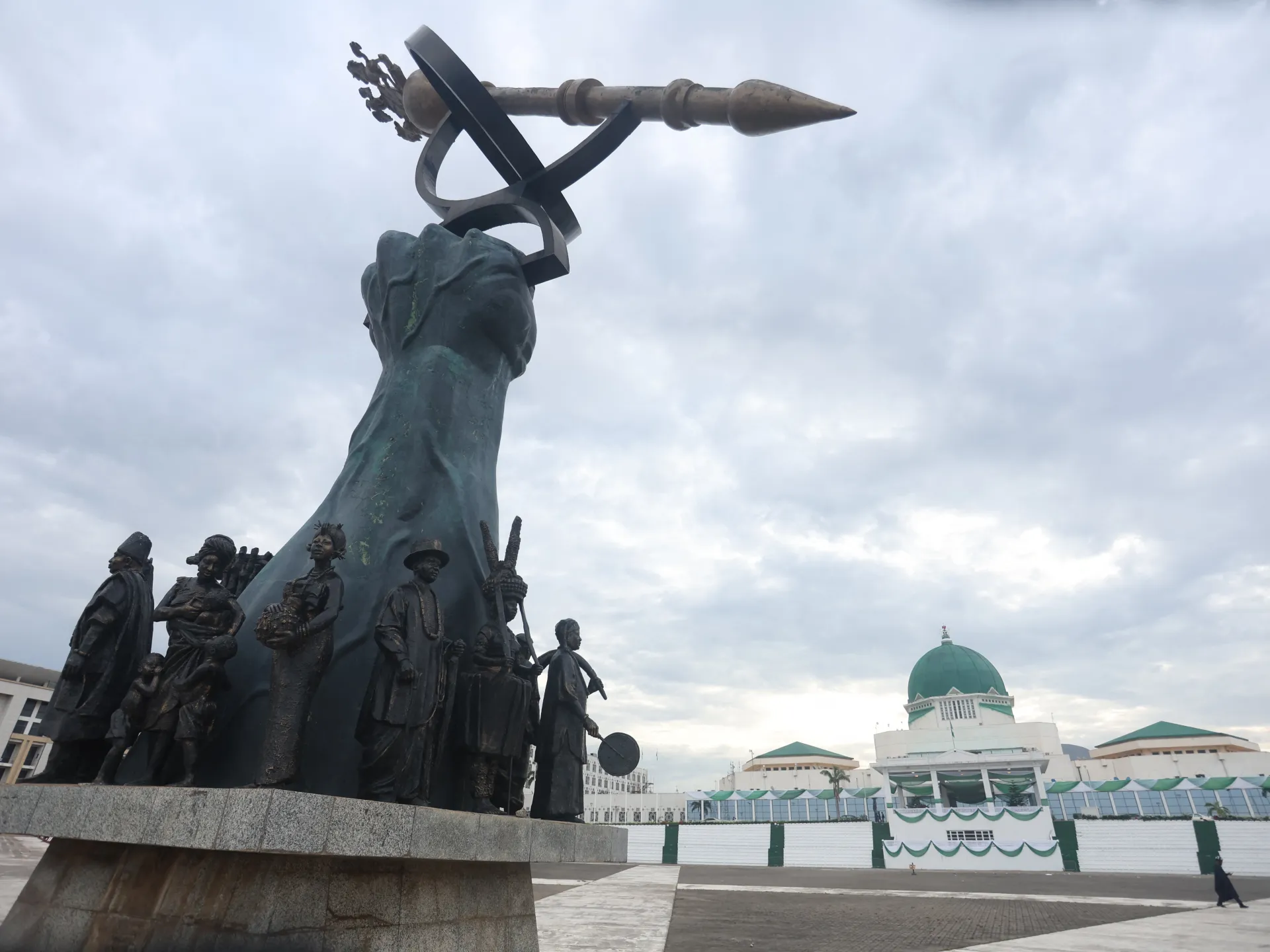 Large historical monument with bronze figures stands before a white building with green domes under cloudy skies.