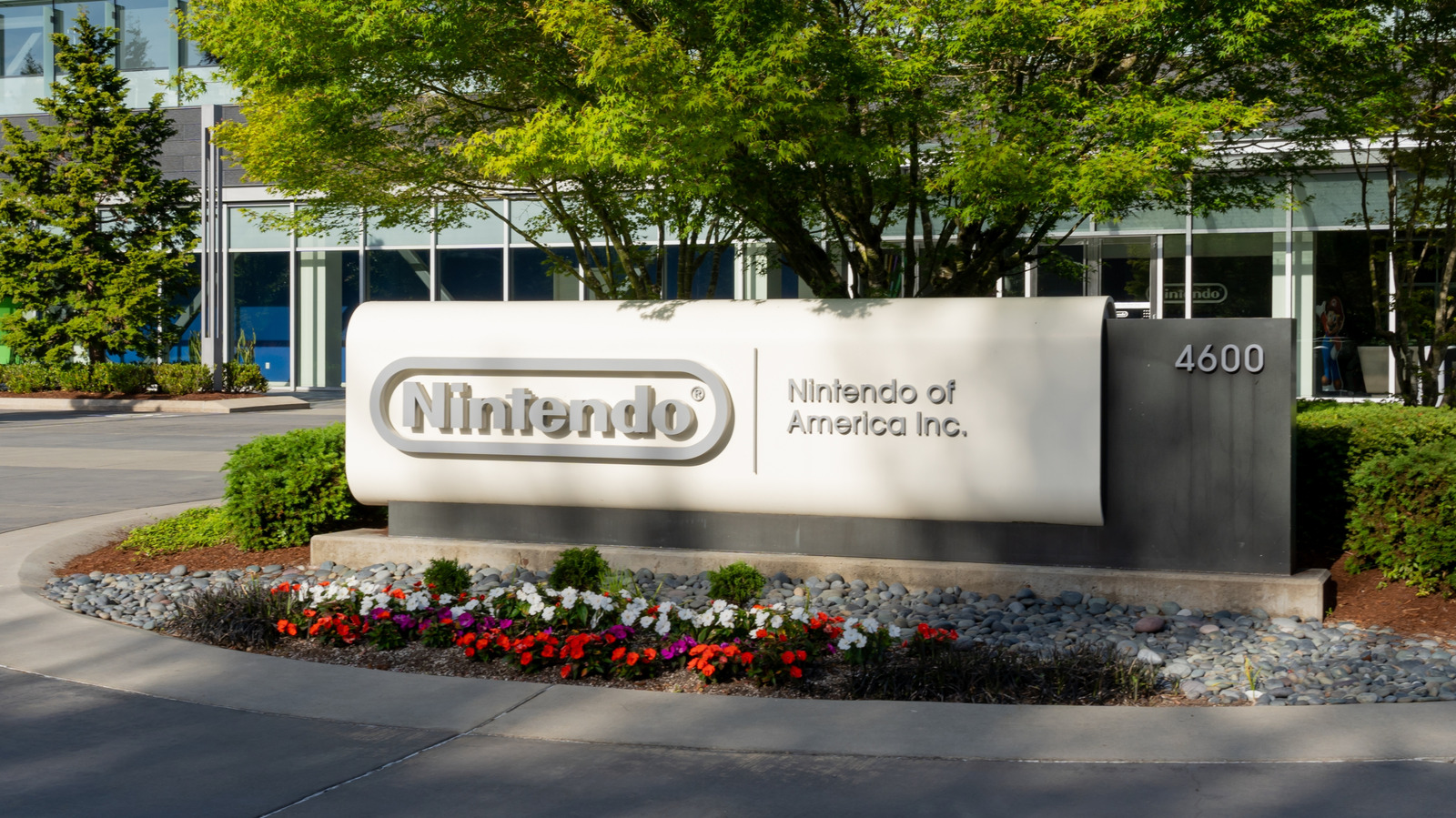 A Nintendo of America sign stands in front of a glass office building, trees, and flower beds.