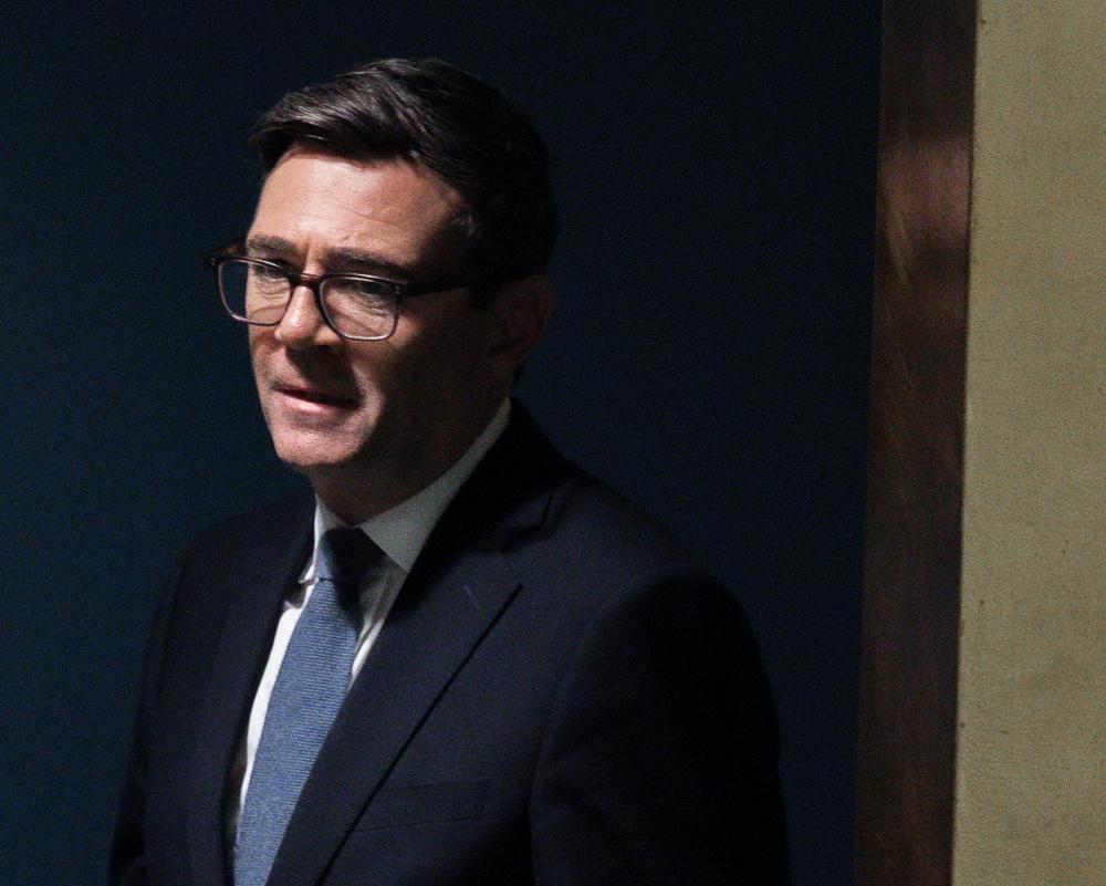 Man wearing glasses, dark suit, white shirt and blue tie against a dark background.
