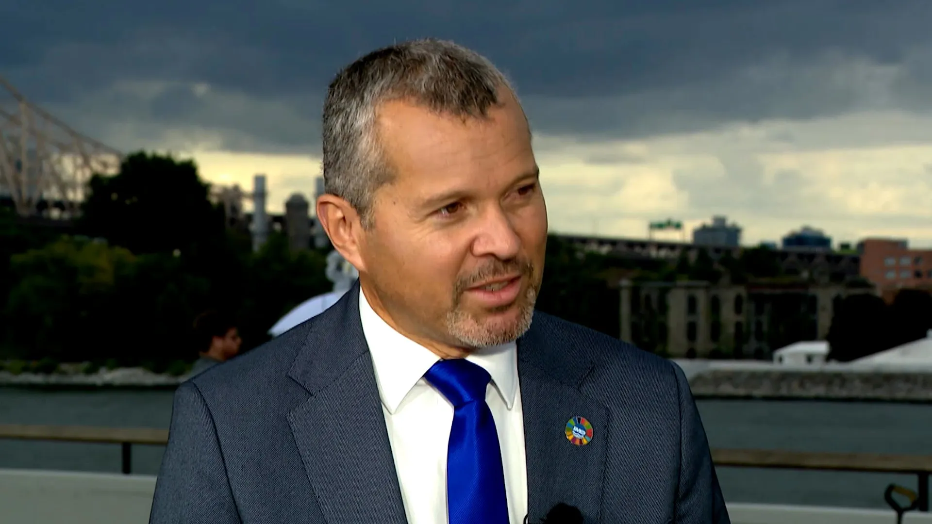A gray-haired man in a navy suit and blue tie speaks in front of a waterfront skyline.