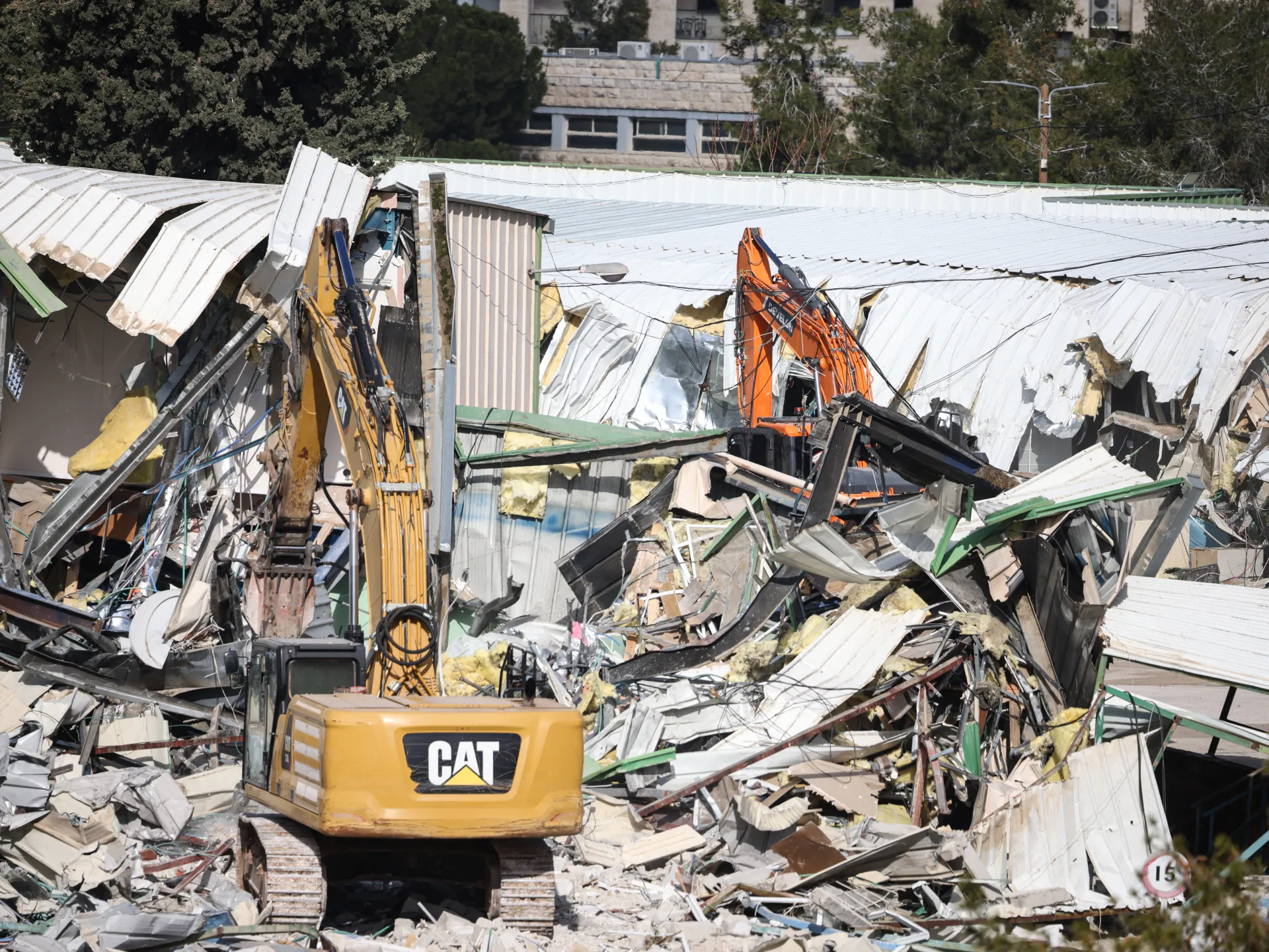 Excavators stand amid the wreckage of a collapsed metal-roofed structure, with debris filling the foreground.