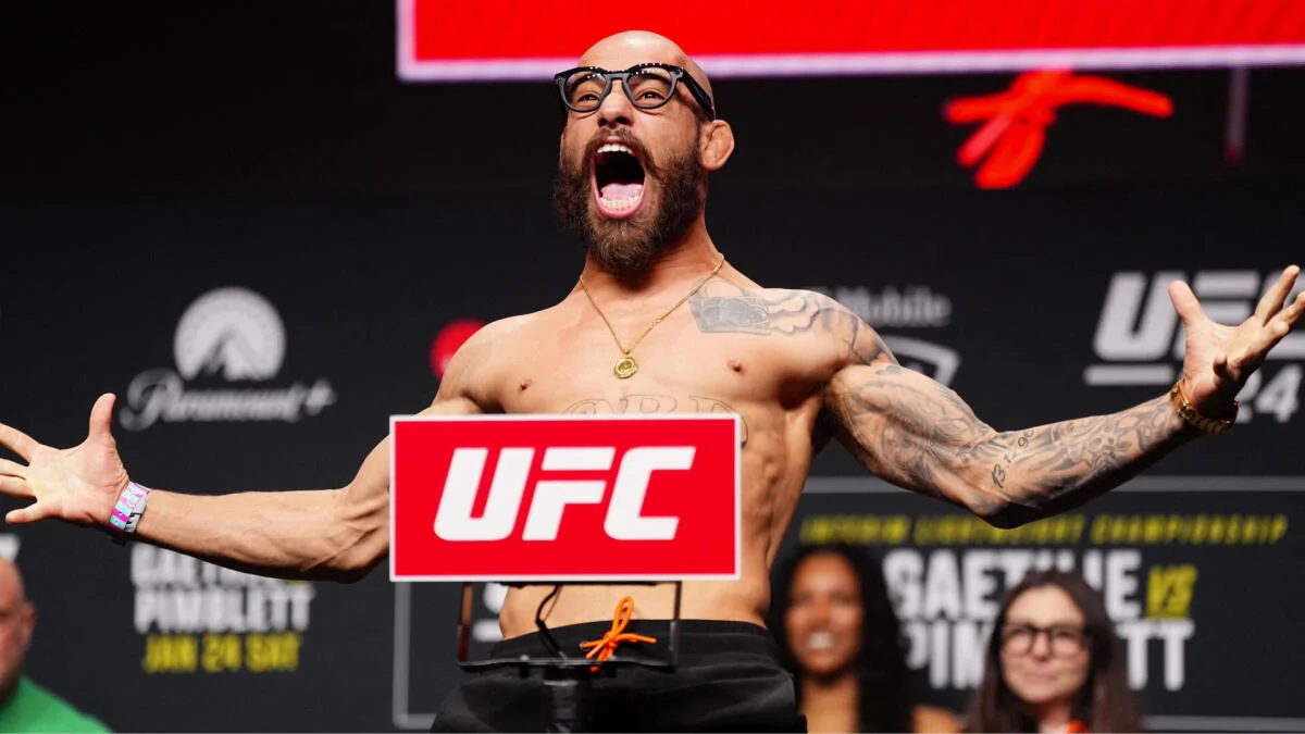 Bearded shirtless fighter wearing glasses spreads his arms beside a UFC sign during a weigh-in.