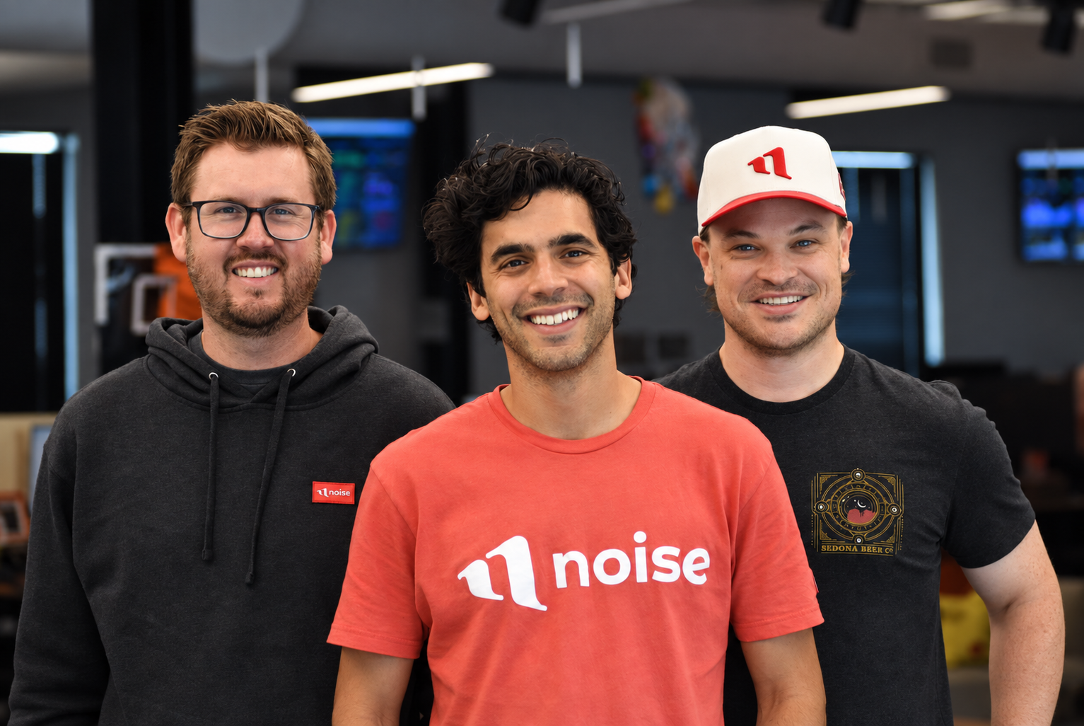 Three smiling men pose together indoors, wearing branded Noise clothing and a baseball cap.