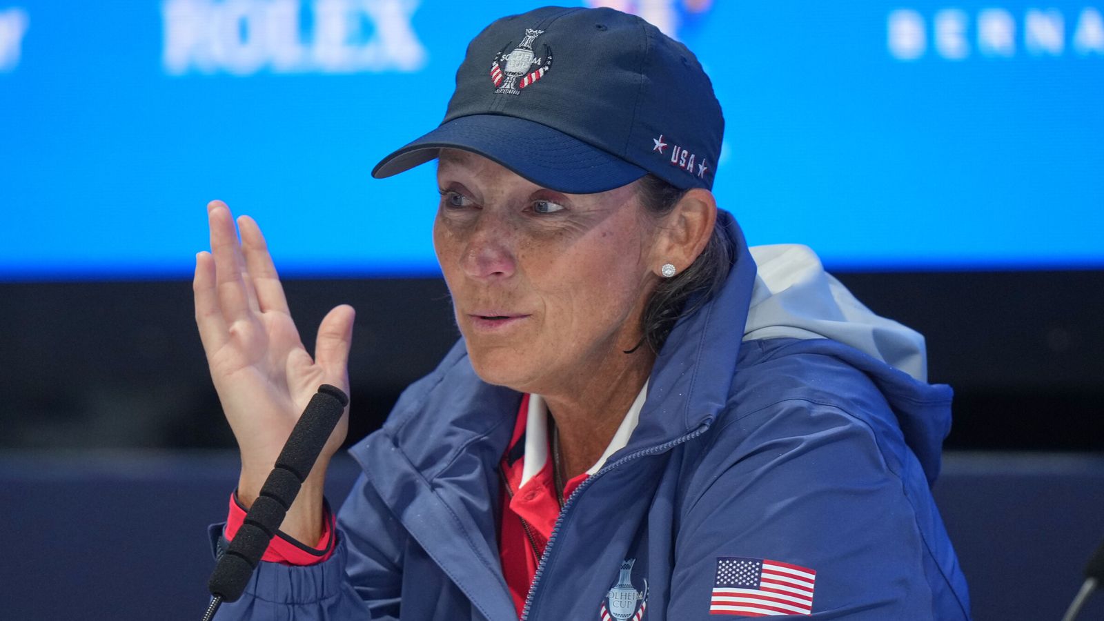 Woman in a USA jacket and cap gestures while speaking into a microphone.