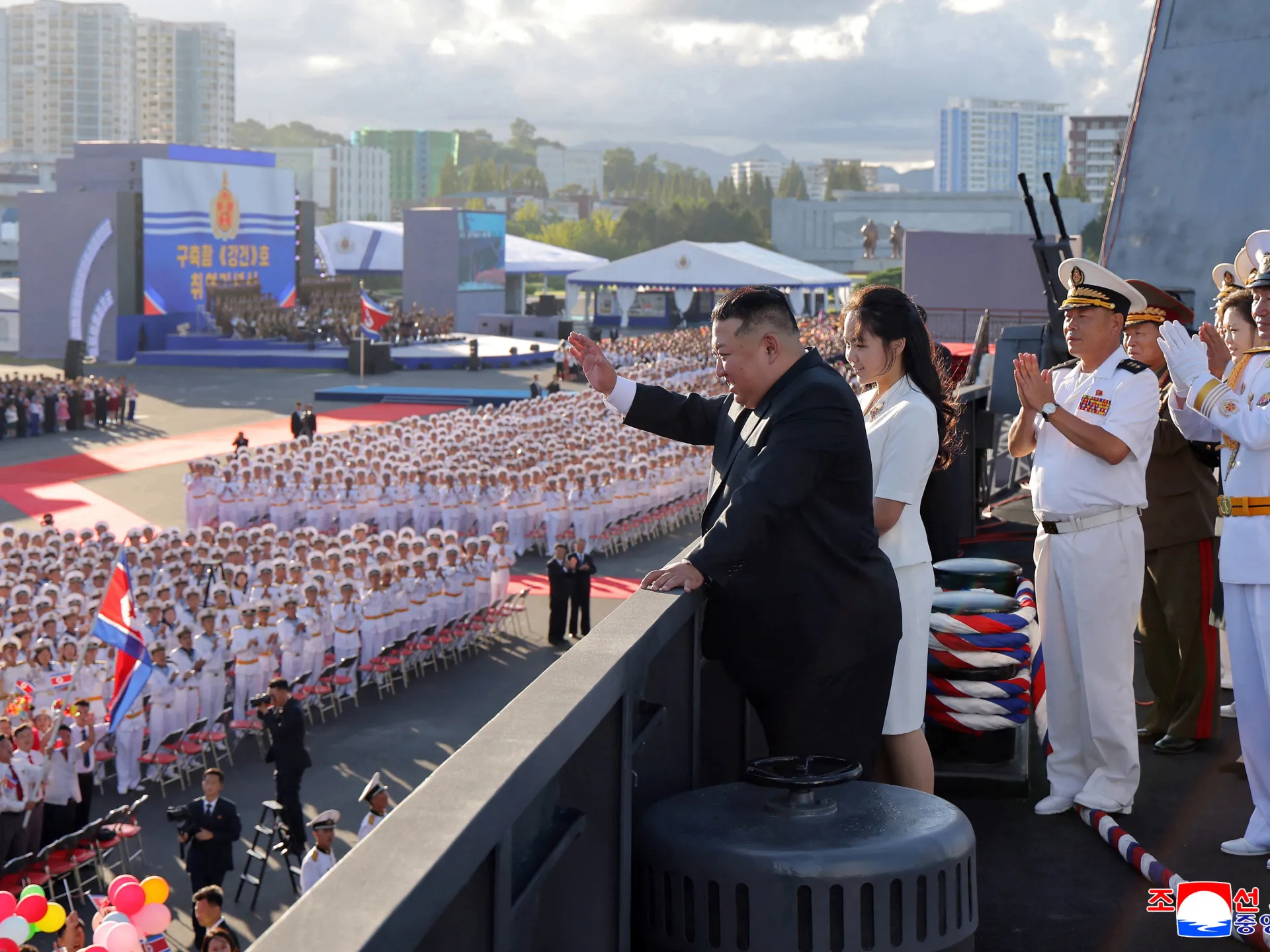 Man in black suit waves from a viewing platform during a large military ceremony