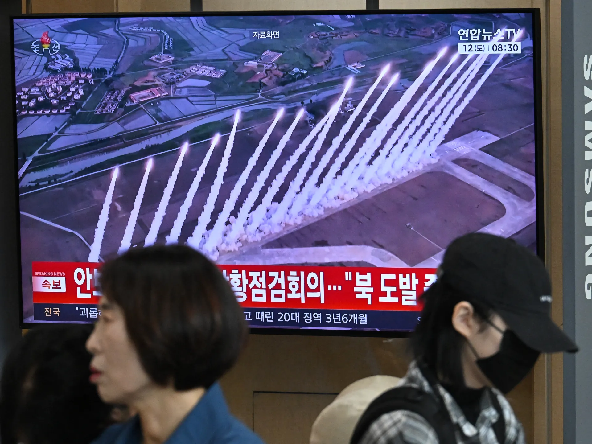 Television displays multiple missile launches over farmland as people pass in front of the screen.