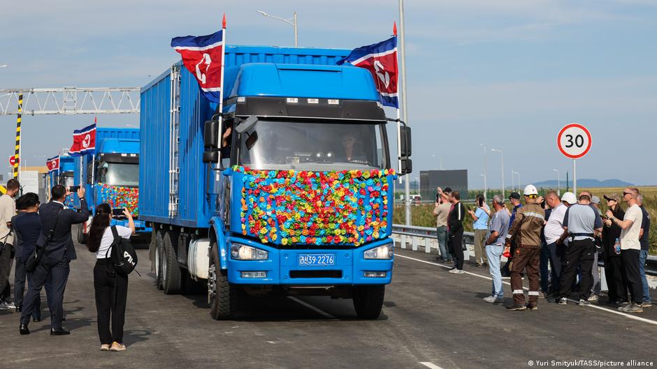 Decorated blue trucks with North Korean flags drive past onlookers along a newly paved road.